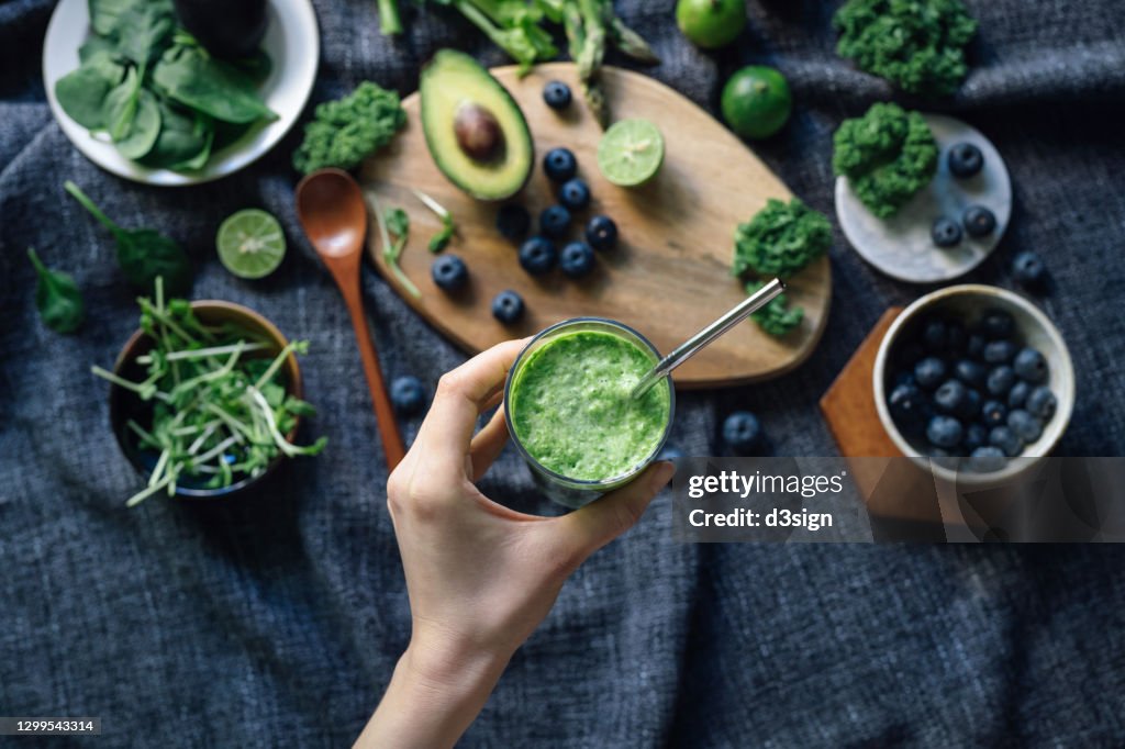 Flat lay of fresh and healthy green fruits and vegetables on a rustic wooden board, against dark grey fabric for a healthy eating diet. Woman's hand holding a glass of green smoothie with reusable straw. Healthy superfood, green colour, detox diet concept