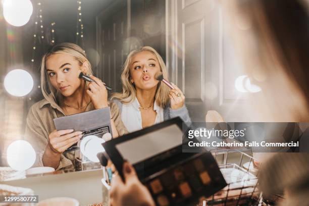 two young woman sit in front of an illuminated mirror and apply make-up - trucco per il viso foto e immagini stock
