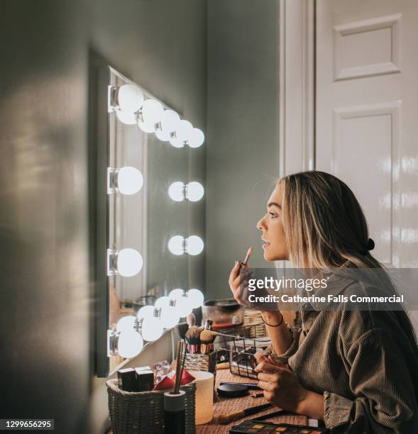 a woman stares into an illuminated mirror as she concentrates while applying lipgloss - vestuario entre bastidores fotografías e imágenes de stock