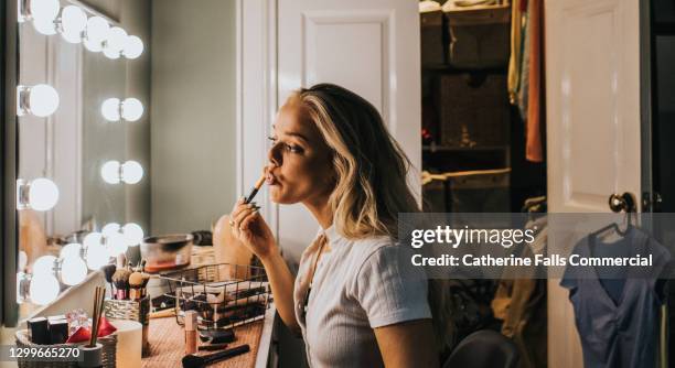 a woman stares into an illuminated mirror as she concentrates while applying lipgloss - woman applying blush stock pictures, royalty-free photos & images