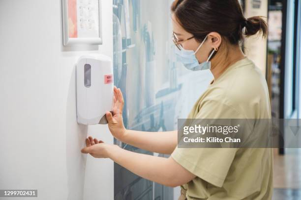 asian woman wearing face mask cleaning her hand with hand sanitizer in an office setting - soap dispenser stock pictures, royalty-free photos & images