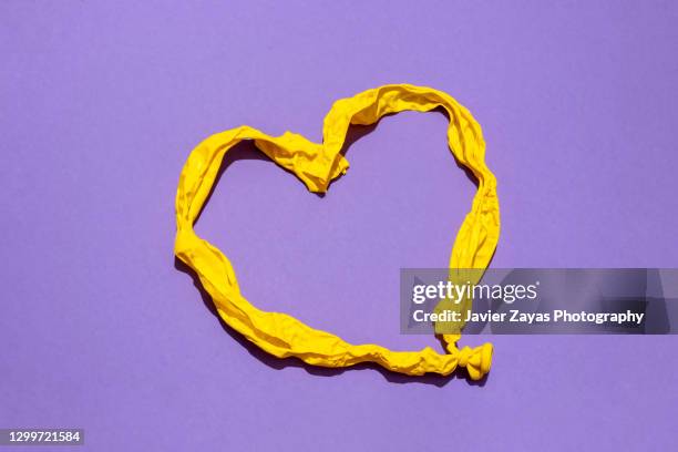 heart shaped deflated yellow balloon against purple background - balloon knot stock pictures, royalty-free photos & images