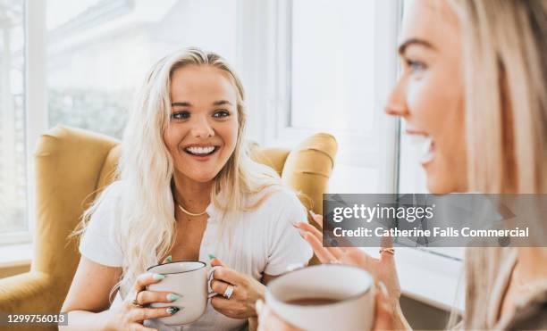 two young woman holding cups of tea. one looks directly at the other and smiles, while she explains something, using her hands to gesture. conversation is lighthearted. - tea leaf stock pictures, royalty-free photos & images