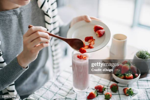 close up of young asian woman making fresh and healthy strawberry smoothie for breakfast, adding chopped strawberries on top as garnish. healthy eating and healthy lifestyle - smoothie stock pictures, royalty-free photos & images