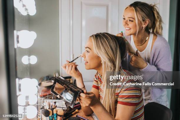 two young woman getting ready - one puts on eyeshadow and the other helps her to do her hair - one friend helping two other imagens e fotografias de stock