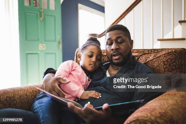 father and daughter reading book in living room at home - lezen stockfoto's en -beelden