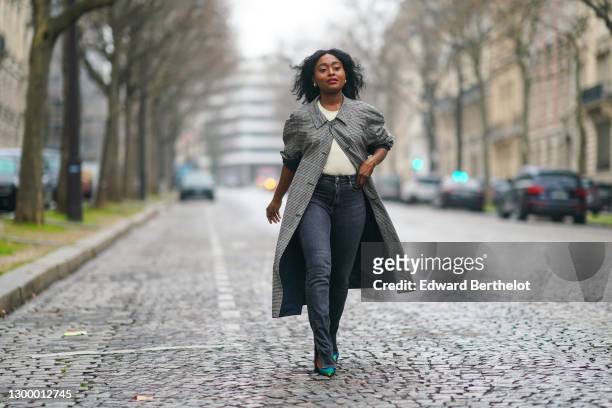 Carrole Sagba aka Linaose wears a gray and black checked long trench vintage coat, a t-shirt from Zara, black denim jeans pants from Zara, blue shiny...
