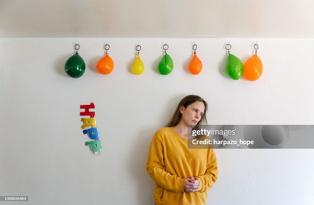 Sad woman standing under deflated balloons beside a broken "happy birthday" sign.