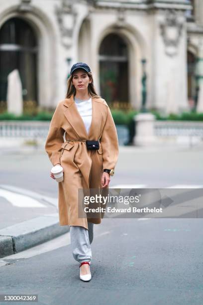 Natalia Verza aka Mascarada wears a black cap from Nike, a white t-shirt, a beige pale brown wool long trench coat, gray sport jogger pants, a golden...