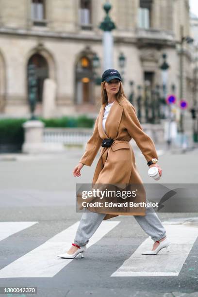 Natalia Verza aka Mascarada wears a black cap from Nike, a white t-shirt, a beige pale brown wool long trench coat, gray sport jogger pants, a golden...