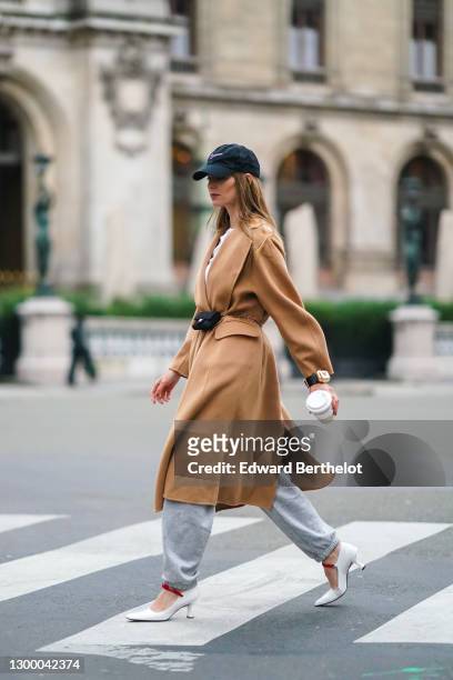 Natalia Verza aka Mascarada wears a black cap from Nike, a white t-shirt, a beige pale brown wool long trench coat, gray sport jogger pants, a golden...