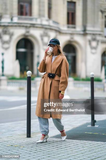 Natalia Verza aka Mascarada wears a black cap from Nike, a white t-shirt, a beige pale brown wool long trench coat, gray sport jogger pants, a golden...