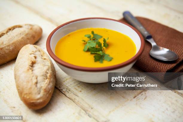 sopa de zanahoria y cilantro en un tazón, cubierta con hojas de cilantro, con panecillos y una servilleta y cuchara marrones, sobre una superficie de madera áspera - semilla de coriandro fotografías e imágenes de stock