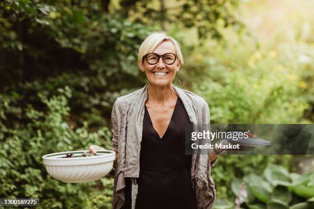 smiling mature woman holding food in bowl and plate against plants in front yard - vrouw 50 jaar stockfoto's en -beelden