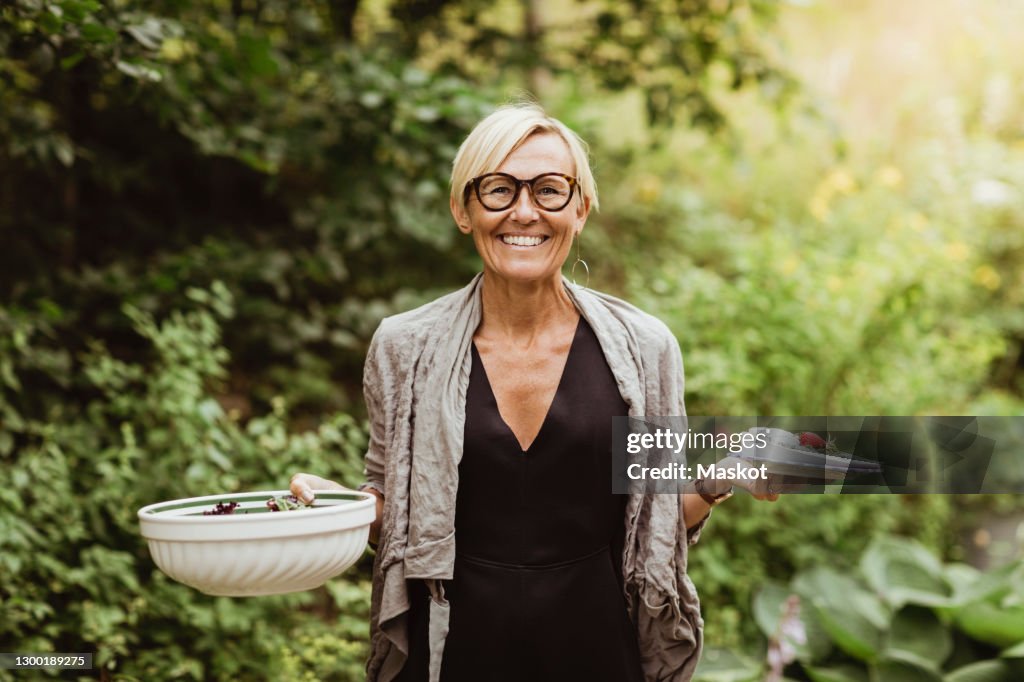 Smiling mature woman holding food in bowl and plate against plants in front yard