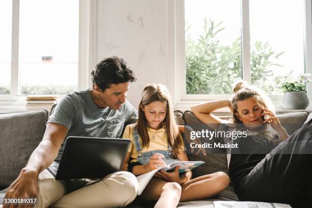 daughter doing homework sitting by mother and father in living room - familie laptop stockfoto's en -beelden