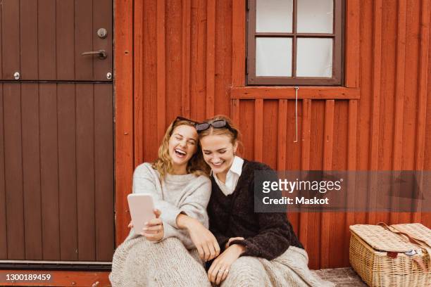 female friends laughing while taking selfie on mobile phone against cottage - schweden stock-fotos und bilder