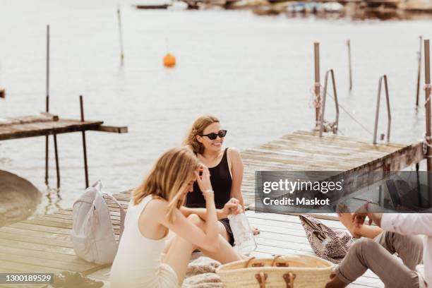 happy young friends sitting on jetty against sea - scandinavia picnic stock pictures, royalty-free photos & images