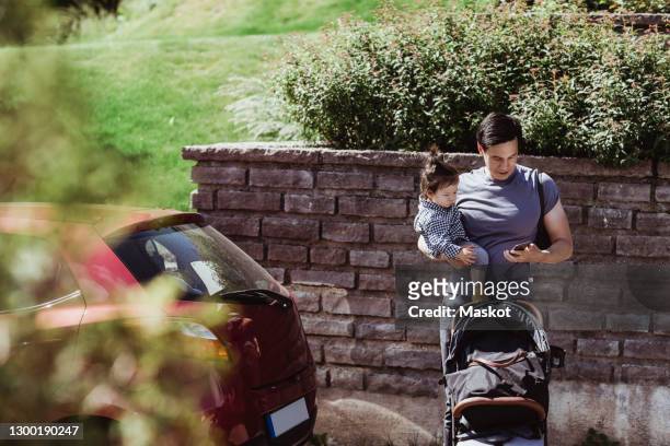 father using smart phone while carrying baby son on sunny day - prams stock pictures, royalty-free photos & images