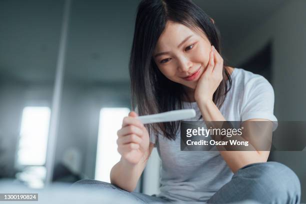 young woman holding home pregnancy test and looking happy in bedroom - asian woman pregnant stockfoto's en -beelden