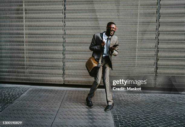 businessman laughing while standing with smart phone on walkway against metal grate - metallic blazer stock pictures, royalty-free photos & images