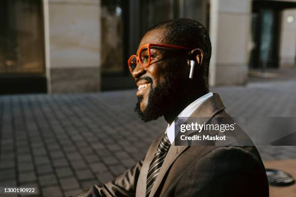 smiling businessman with eyeglasses looking way while sitting on footpath - formelle geschäftskleidung stock-fotos und bilder