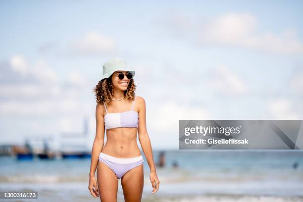 smiling young woman in a sun hat and bikini walking on a beach - girls swimwear stock pictures, royalty-free photos & images