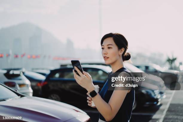 confident and professional young asian businesswoman walking to her car in an outdoor carpark in the city, using smartphone and holding a cup of coffee. business on the go concept - buying a car 個照片及圖片檔