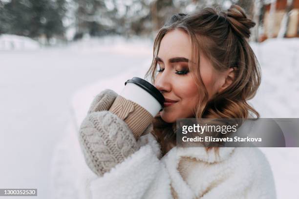 portrait of a young beautiful cute woman with a curly hairstyle warms herself and drinks coffee while holding a glass with her hands in mittens in a snowy forest - woman hands in mittens stock-fotos und bilder