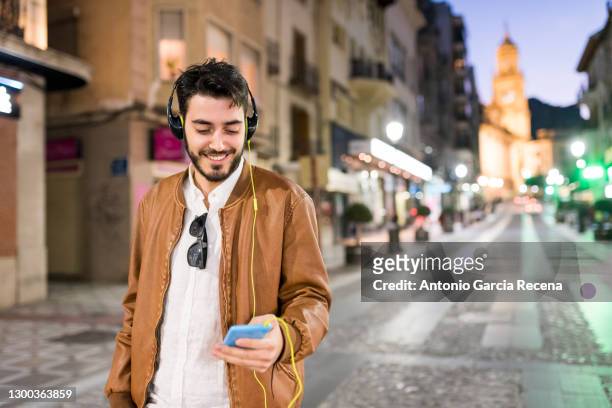 happy causal man looking smart phone smiling in city night image. listening musci with headphones - jaén city stock pictures, royalty-free photos & images