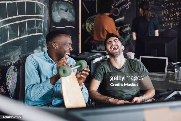 cheerful it professionals laughing while sitting with skateboard at office - lachen stock-fotos und bilder