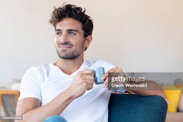 smiling man drinking coffee while sitting on chair at home - espresso trinken stock-fotos und bilder