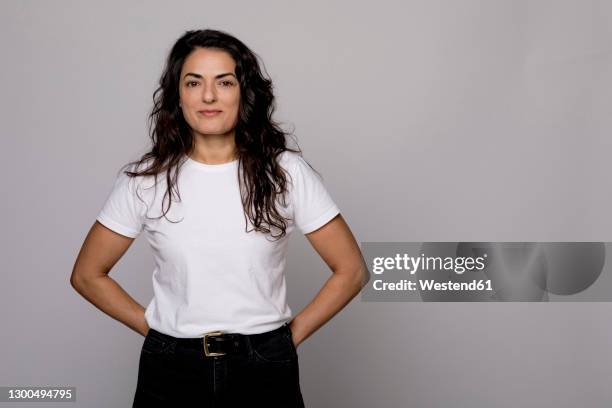 beautiful smiling woman standing with hands behind back against gray background - white t shirt stockfoto's en -beelden