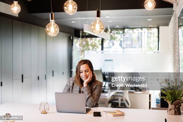 smiling businesswoman with hand on chin using laptop at desk in office - smartphones dangling stock pictures, royalty-free photos & images