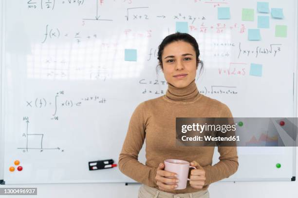 female student holding coffee cup while standing against whiteboard at home - women in stem foto e immagini stock