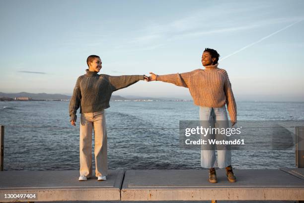 friend stretching hands while standing at distant on bench against sea - far stock pictures, royalty-free photos & images