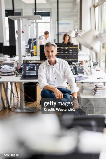 mature businessman sitting on chair with colleague standing in background at open plan office - open day 3 stock-fotos und bilder