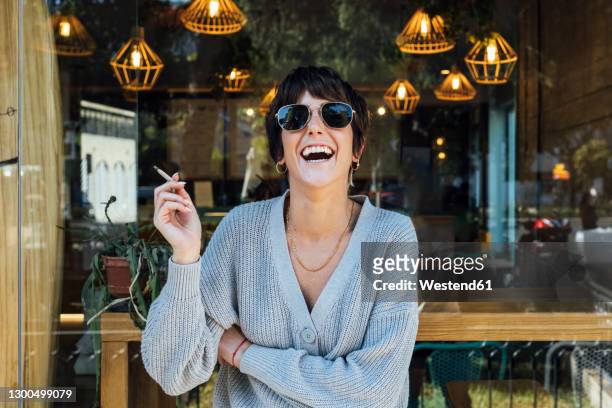 young woman wearing sunglasses smoking cigarette while standing against restaurant window - thema rauchen stock-fotos und bilder