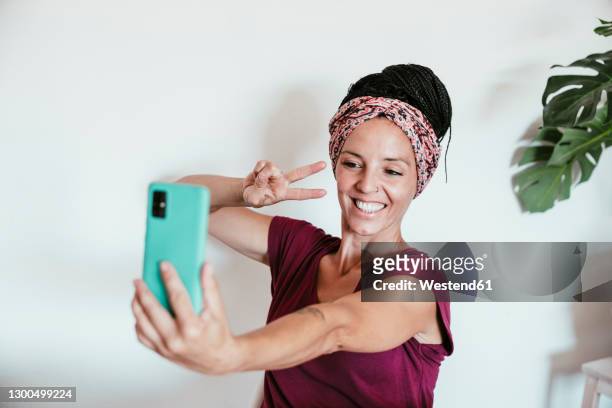 smiling woman taking selfie on smart phone against wall at home - mid adult women photos et images de collection