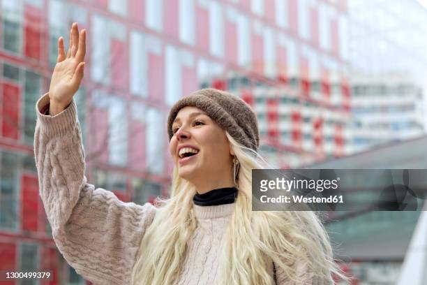 cheerful young woman waving hand while standing against building in city - sventolare la mano foto e immagini stock
