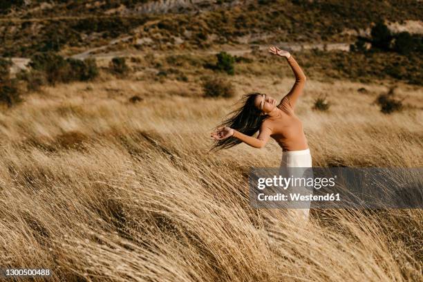 young woman with arms raised standing amidst grass in field during windy day - flatterndes haar stock-fotos und bilder