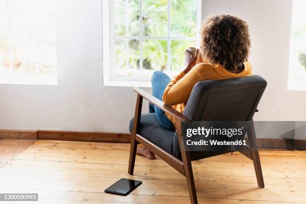 woman drinking tea while sitting on armchair at home - behind window stock-fotos und bilder