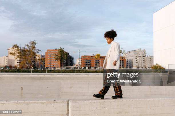young woman with afro hair walking on retaining wall against buildings in city - balancieren mauer stock-fotos und bilder