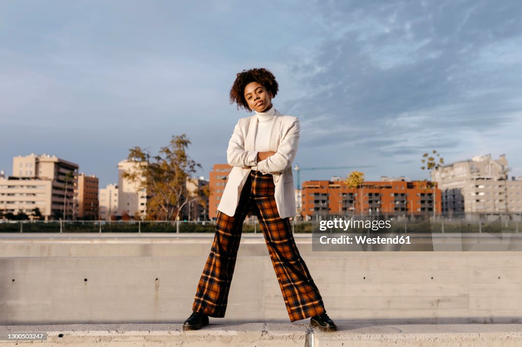 Carefree afro woman with arms crossed posing on retaining wall against buildings in city
