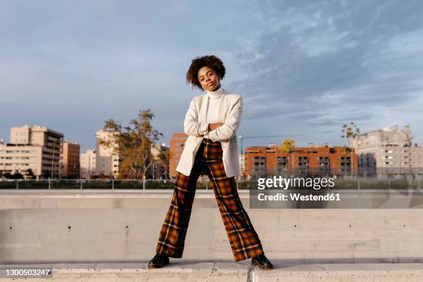 carefree afro woman with arms crossed posing on retaining wall against buildings in city - fashion stock-fotos und bilder