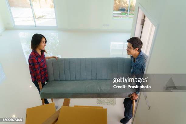 happy young asian couple lifting sofa or couch into living room with cardboard box at their new home together. moving house day. - carrying sofa stock pictures, royalty-free photos & images