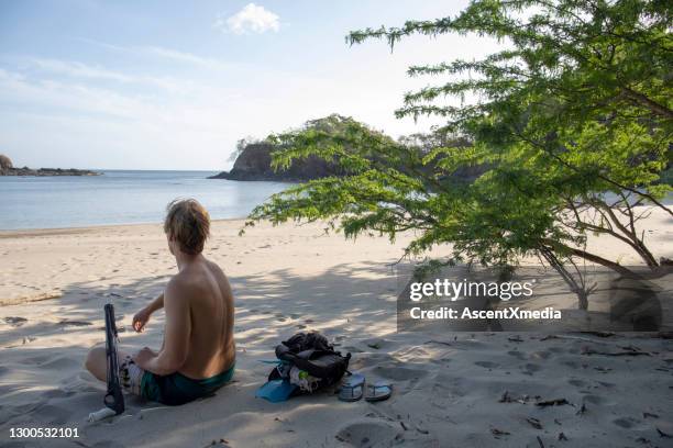 young man with speargun relaxes on white sand beach in the morning - man spear fishing stock pictures, royalty-free photos & images
