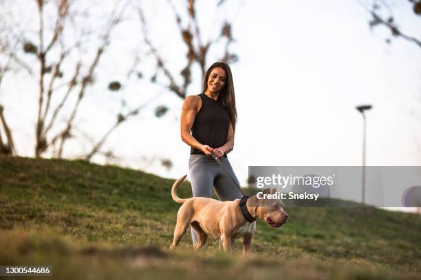 smiling young lady, muscular-built, walking her stafford in park. - stafford terrier stock pictures, royalty-free photos & images