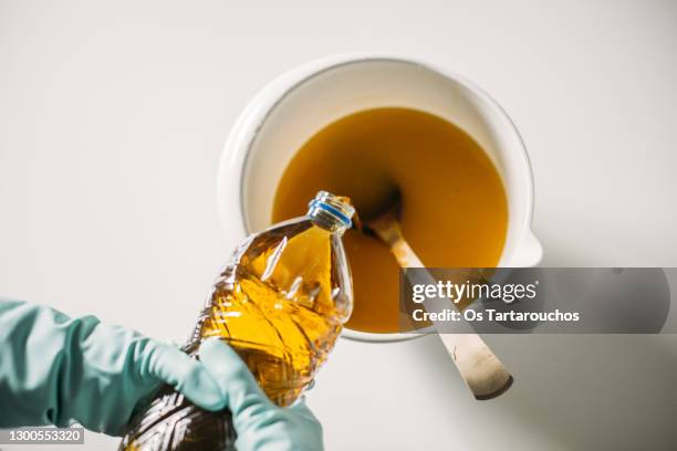 soap making at home - hands wearing protective rubber gloves pouring oil into a bowl with water and sodium hydroxide mixture in a white pastic bowl with a wooden spoon - handschuhe stock-fotos und bilder