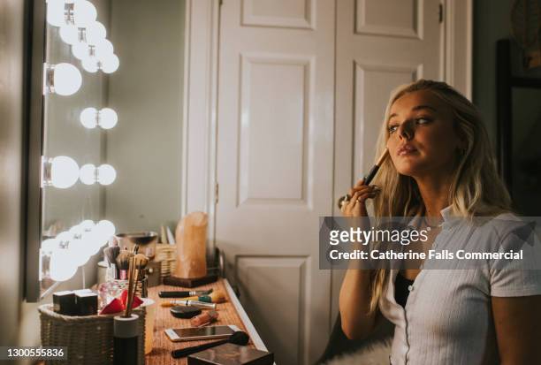 a young woman contours in front of an illuminated mirror - geschminkt gezicht stockfoto's en -beelden